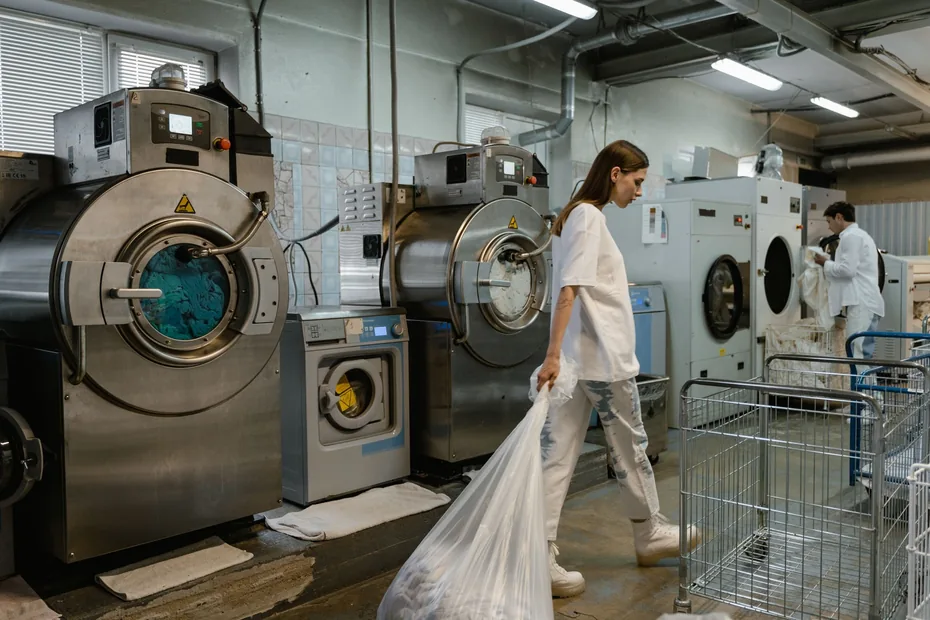 A worker in an industrial laundry, with large washing machines and linen trolleys